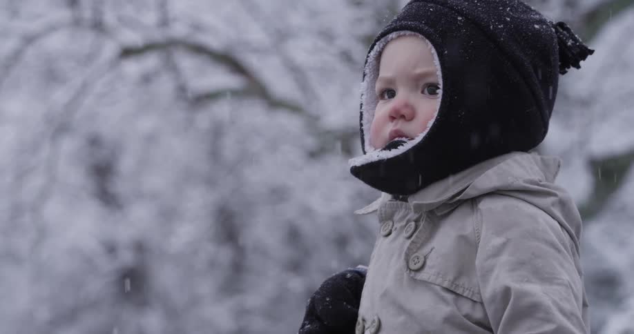 A bundled up toddler walks and lifts his hand to catch snow outside while snow is falling