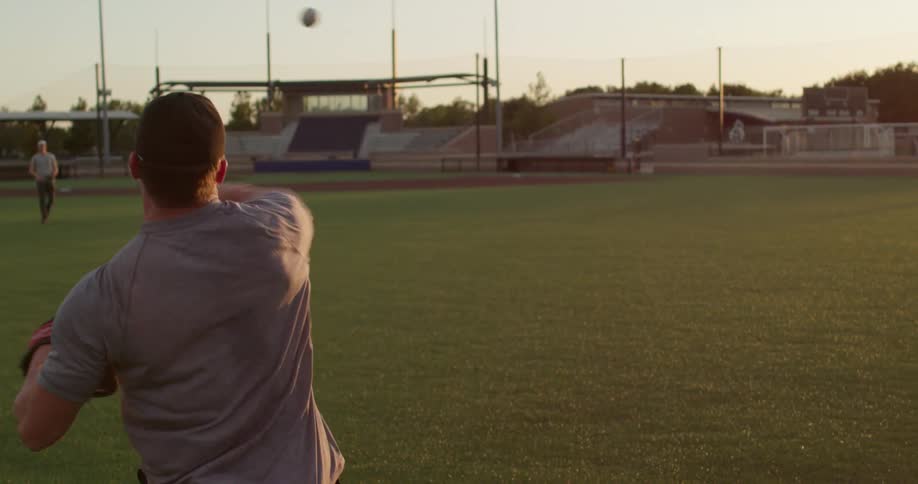 Man pitches a baseball to another man playing on a large baseball field at sunset
