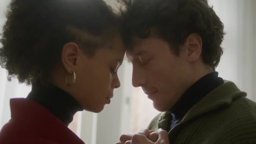 A man and a woman standing in a room with foreheads touching with a window in the background