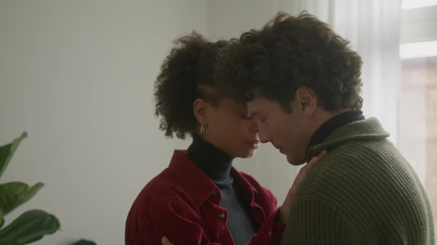 Husband and wife embracing while standing in a living room during the day with their heads pressed up against each other