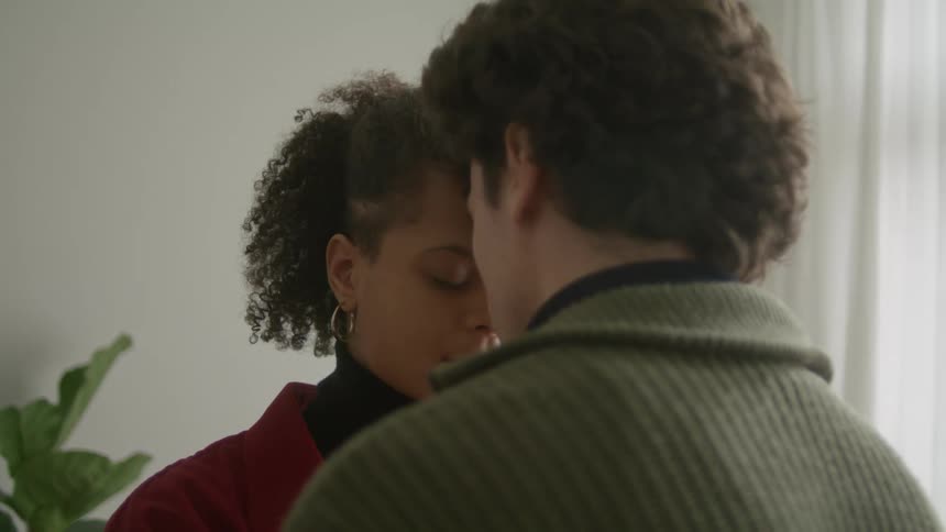 A husband and wife press their heads together and embrace while standing in a living room indoors at daytime