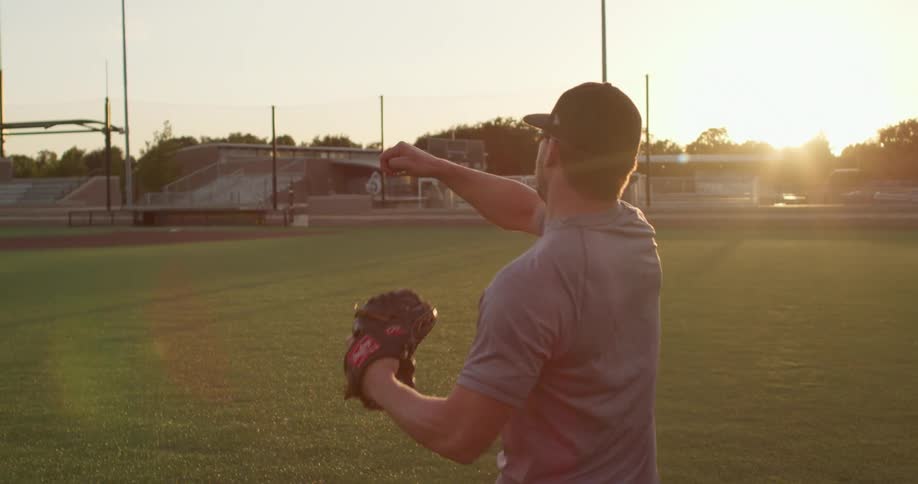 A man wearing a baseball glove pitches his arm back then throws a baseball on a baseball field as a flare of sunlight peers from behind him at sunset