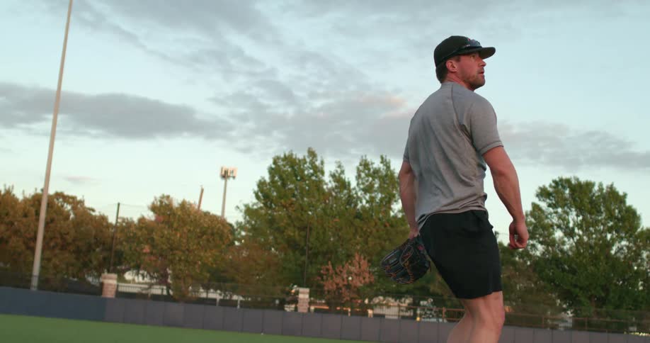 A man in a baseball field throwing a baseball with trees the sky and clouds in the background