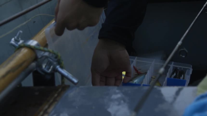 Hands reach into a tackle box on a boat and pick out a lure