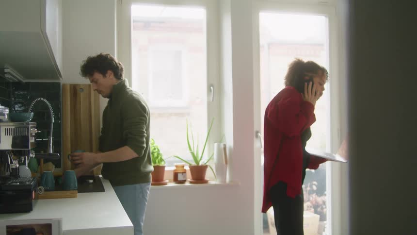 A woman talks on her phone while walking and holding a laptop while her husband washes the dishes