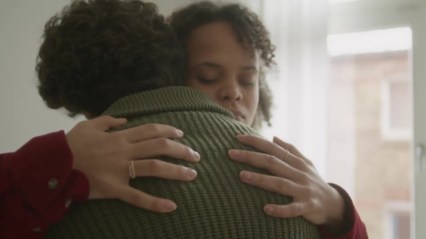 A man and a woman in a room hugging with a wall and window in the background