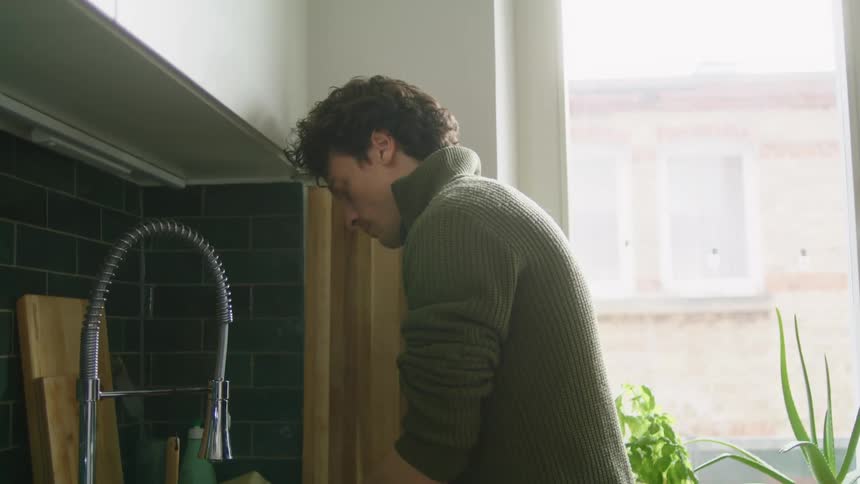 A man stands at the kitchen sink washing mugs as sunlight comes in through the large window