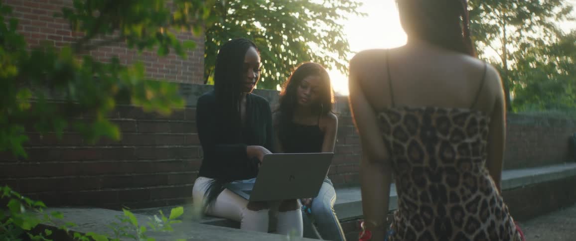 A woman shows her friendly something on her laptop as they sit on a bench outside a brick building
