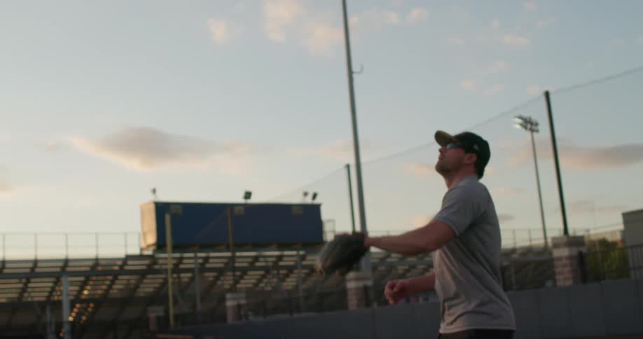 A man catching a baseball with a glove on a stadium at sunset