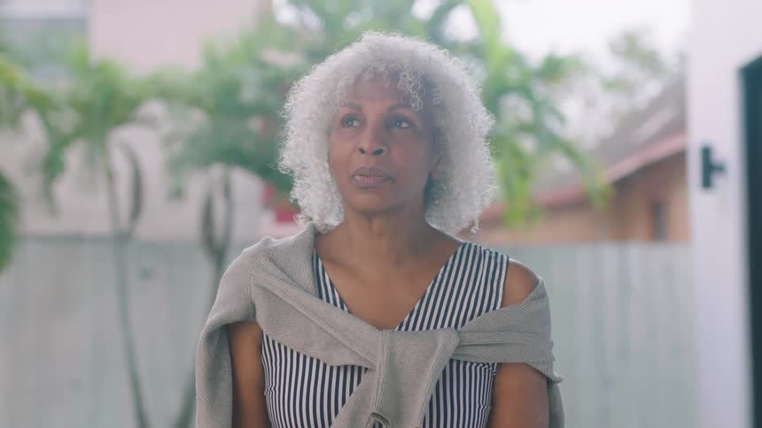 A woman sitting outside at daytime in a backyard closes her eyes and takes a deep breath then opens her eyes and looks around