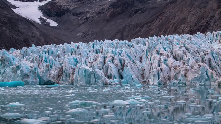 Small pieces of ice moving around on the surface of water next to ice glaciers pointing out from a rocky mountain slope