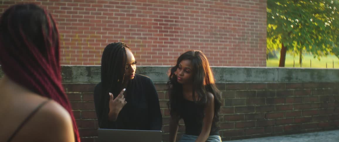 Group of female friens sitting outdoors near a red brick building looking at a laptop at sunset