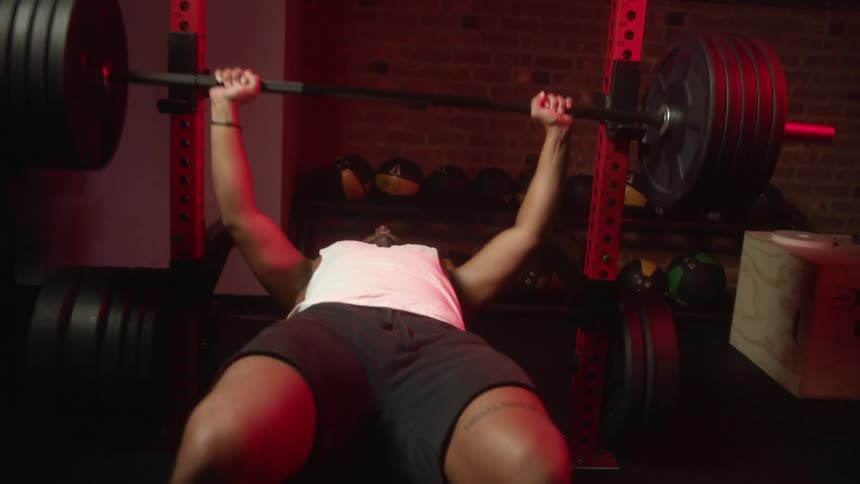 A man in a gym bench pressing weights with a wall and medicine balls in the background