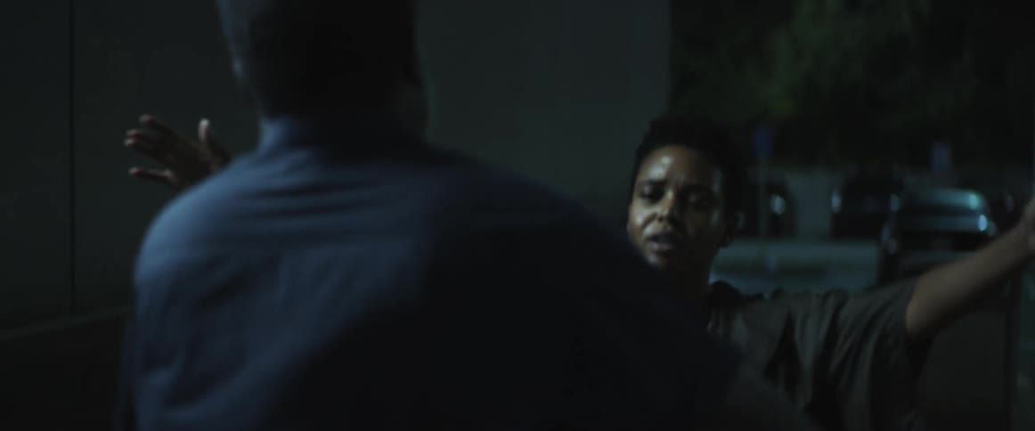 A woman runs over to a man standing outside in a parking lot at night and gives him a hug