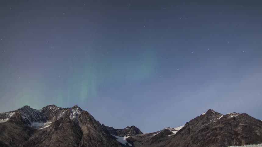 A coast with water and glaciers with a mountain range with the aurora borealis and the sky and clouds above