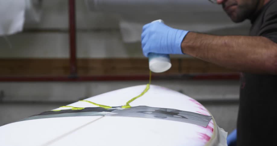 A man pouring a yellow liquid on the surface of a surfboard inside a workshop
