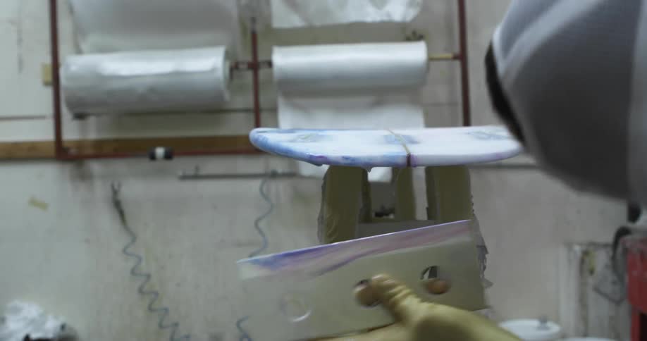A man uses a large plastic blade to scrape and smooth paint on a surfboard in a factory