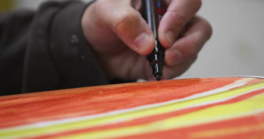 A man drawing a line with a black marker over the surface of a surfboard inside a workshop