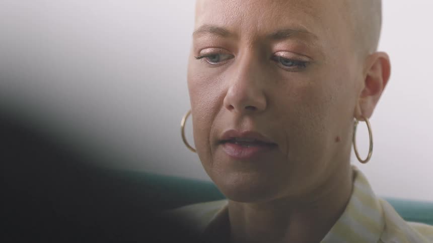 Bald woman with blue eyes wearing metallic earrings talking to someone in a room