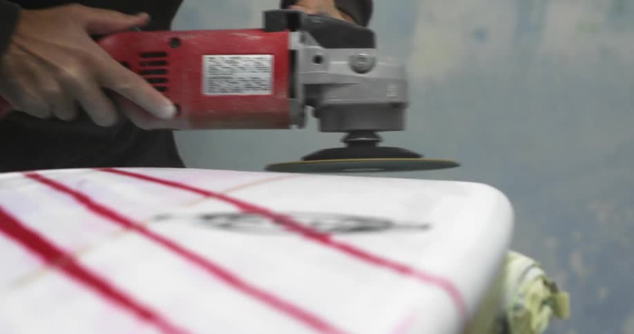 A person in a room sanding a surfboard with an electric sander