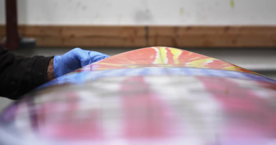 Man wearing a blue disposable glove smoothing a clear liquid on the edges of a surfboard propped up onto a table
