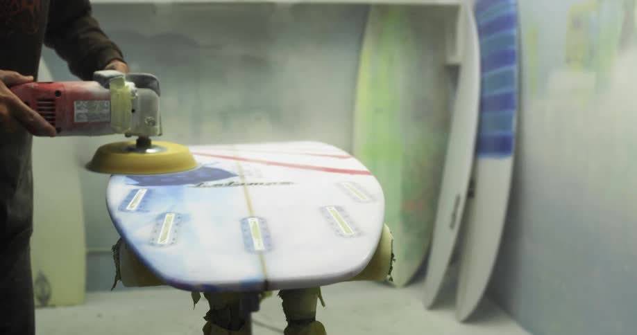 A man in a shop sands a surfboard