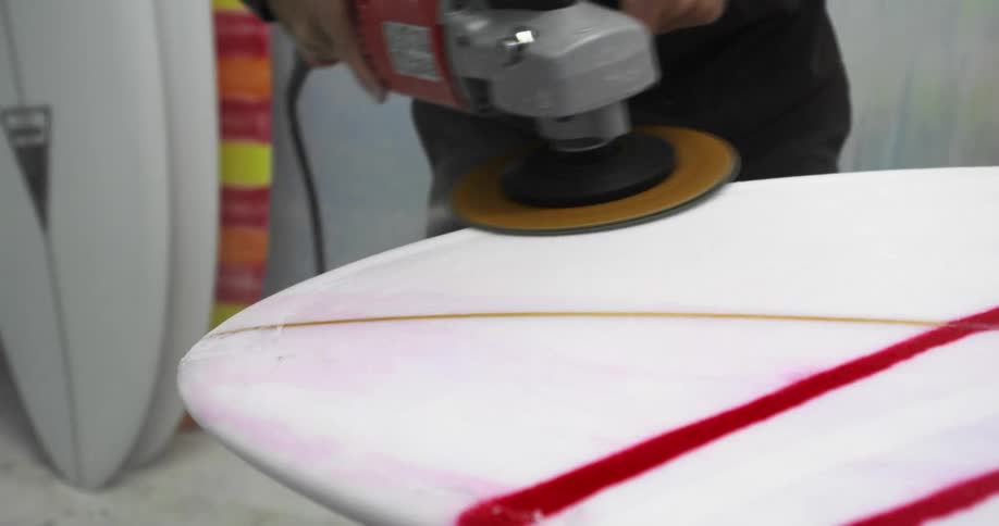Man buffs surfboard with power tool near other surfboards stacked against wall