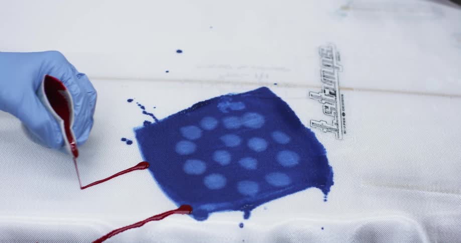 A gloved hand pours red paint from a paper cup in a straight line to add stripes to a flag on a surfboard