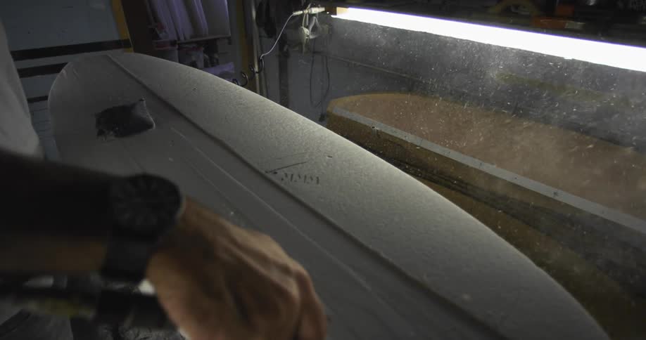 A man uses a tool to wood dust off of a surfboard being crafted in a workshop
