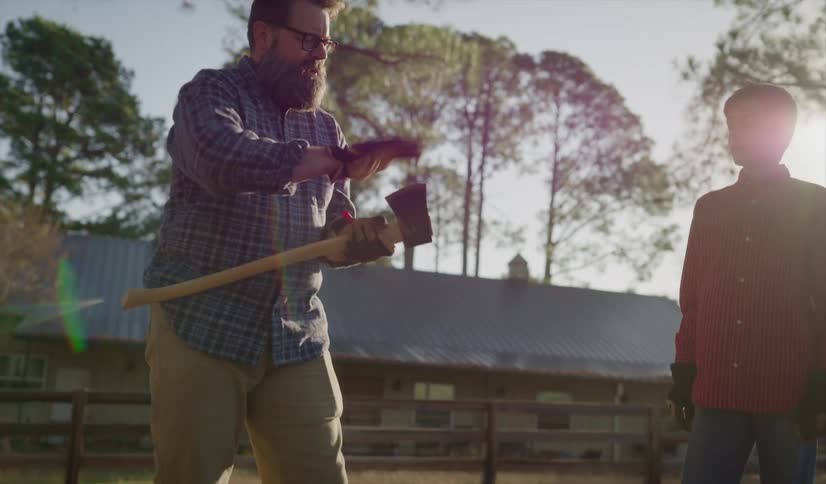 A boy hands an ax to an older man who examines it carefully as the two stand outdoors during the day