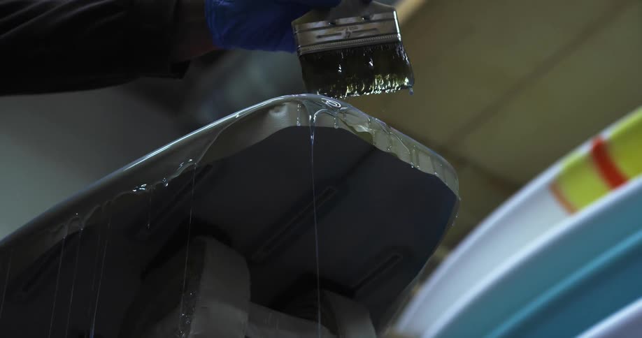 A hand wearing a blue latex glove brushing a paint brush back and forth on the surface of a board while liquid pours off the sides
