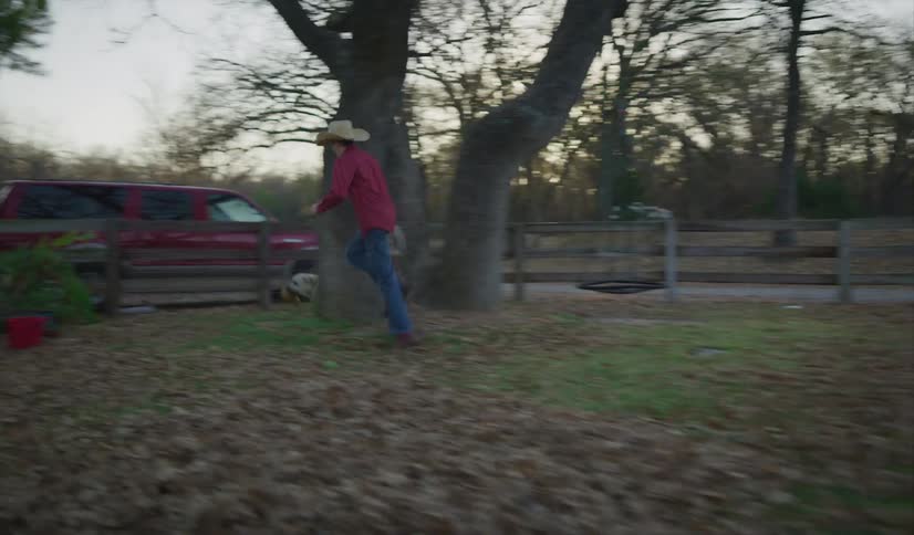 A boy with a cowboy hat chasing a dog in a yard with trees the sky and clouds in the background