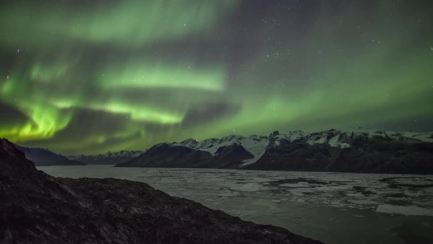 The northern lights move across the sky over a mountain range and ice covered lake