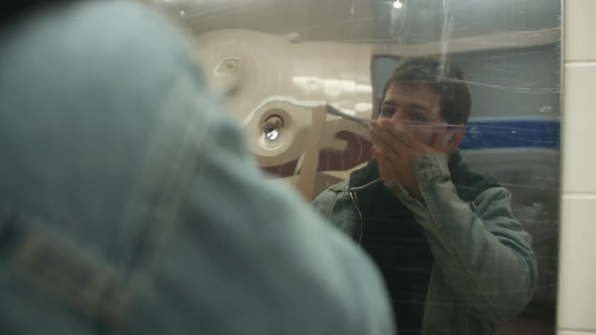 A man with a jacket standing in a bathroom and looking at his reflection on a mirror