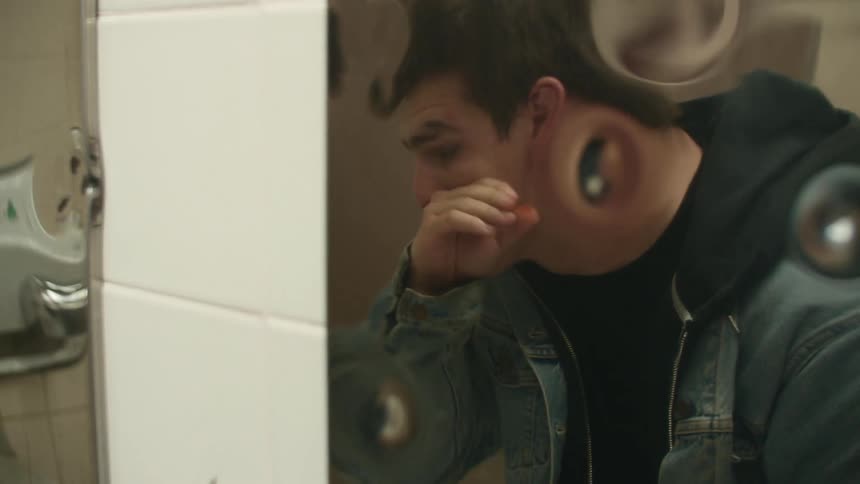 A teenage boy wipes his face as he looks in a mirror in a public bathroom