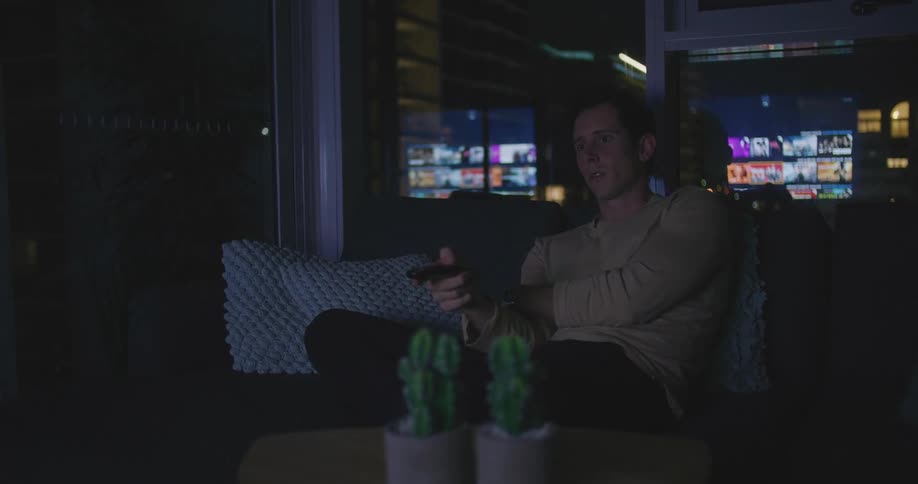 A man uses a remote to navigate the menu on his television as he sits on a sofa in a dark room at night