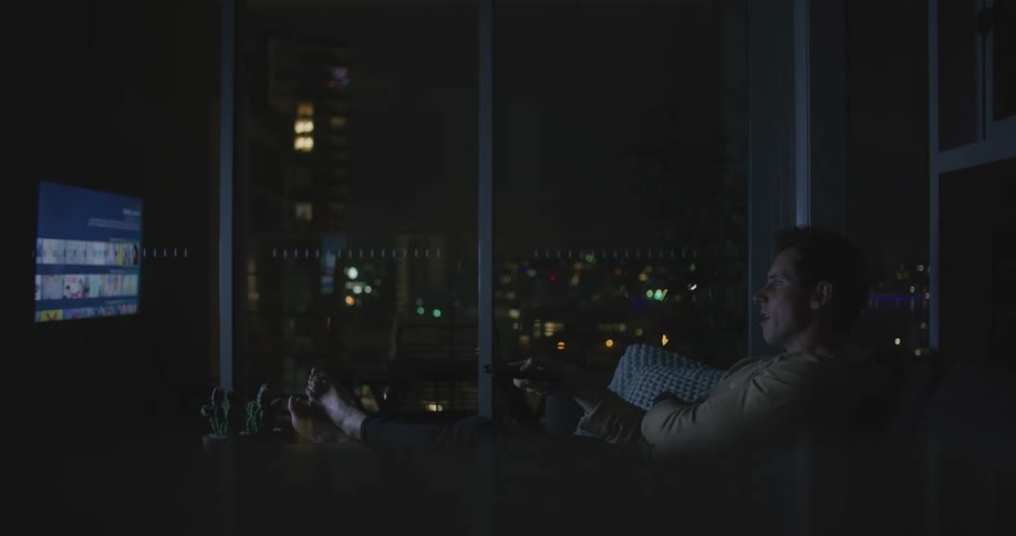 Man with his feet propped up on a coffee table sitting on a sofa scrolling through a tv with a remote control at nighttime