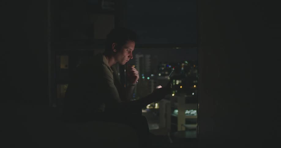 A man sitting in a room texting on his phone at night