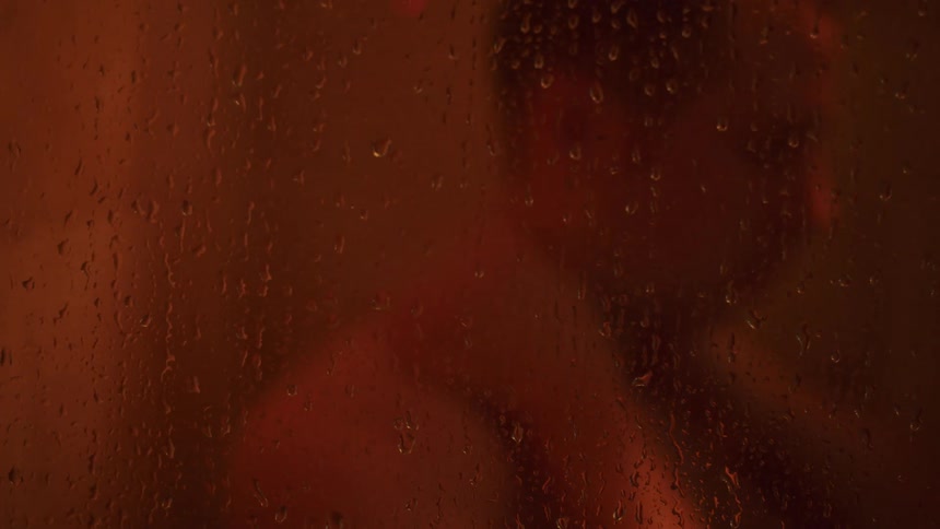 A man behind a shower glass with water droplets who has someone elses arms wrapped around his neck