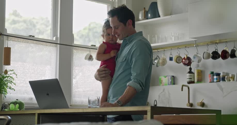Man holding her baby daughter in the kitchen at home talks to someone on a video call on a laptop