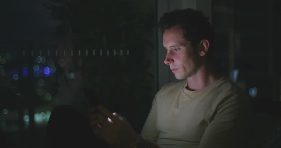 A man uses his thumbs to type on his smartphone while sitting beside a large window in a dark room at night