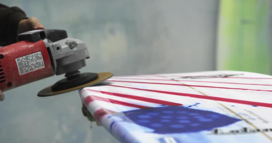 A person with an electric sander sanding a surfboard with a wall in the background