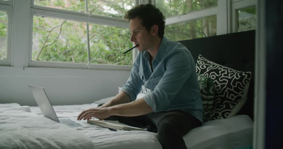A man sits on his bed and types on his laptop and writes in a notebook