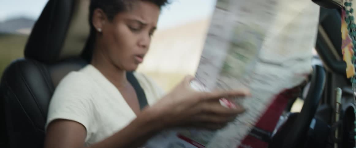 A woman in a vehicle unfolding a map with a rosary on the rearview mirror