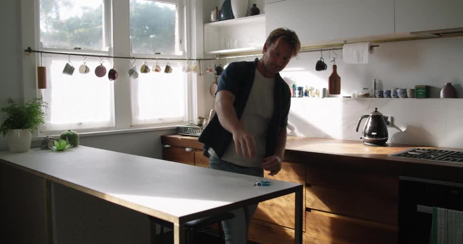 A middle aged man retrieves a large bag and keys from a kitchen counter before walking out of frame