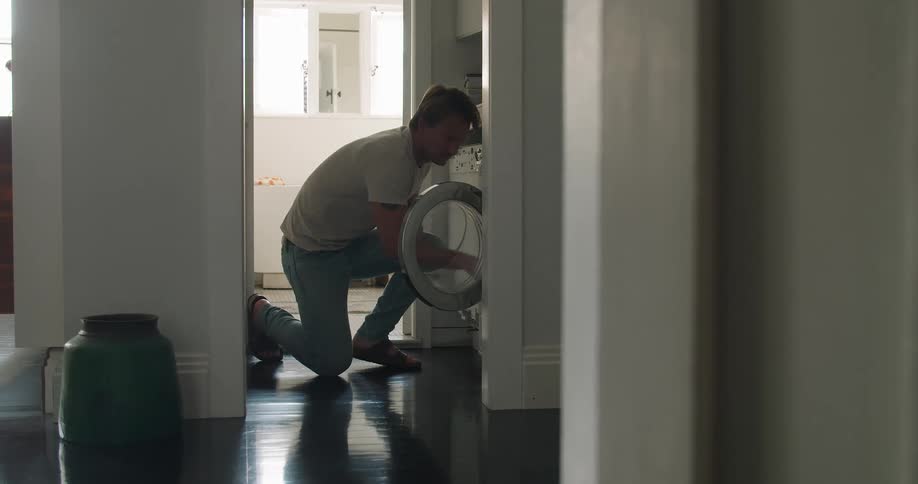 A man opens a front loading washing machine and places a bundle of clothing inside while on one knee inside of a house