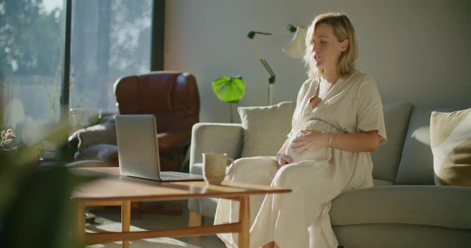 A pregnant woman sitting on a sofa in a living room waves while talking in a video chat on a laptop during the day