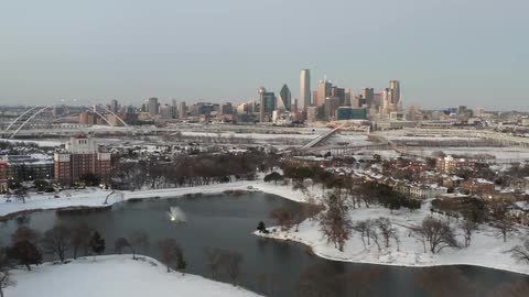 An aerial view of a neighborhood with snow next to a small lake with a distant view of high rise buildings on the horizon