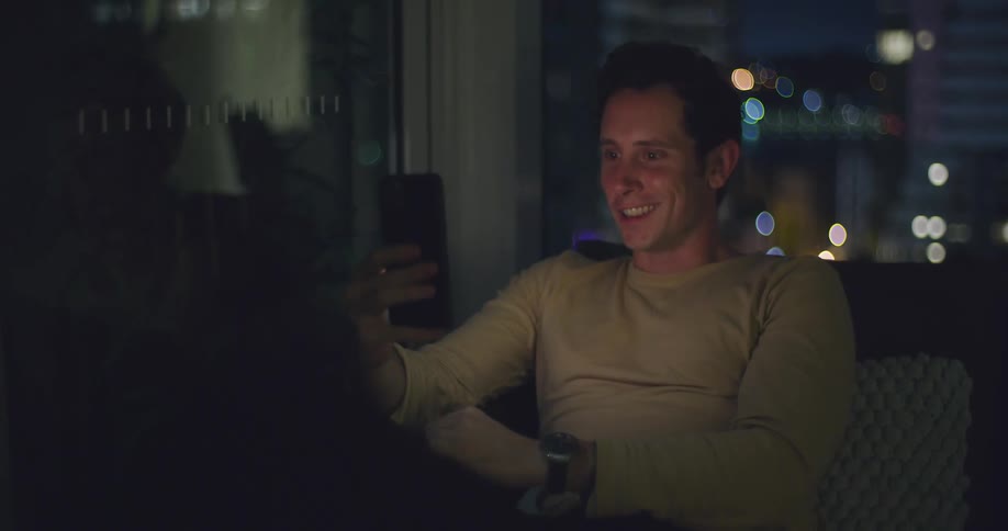 A man sitting in a couch laughing and talking on a video call on his smartphone at night