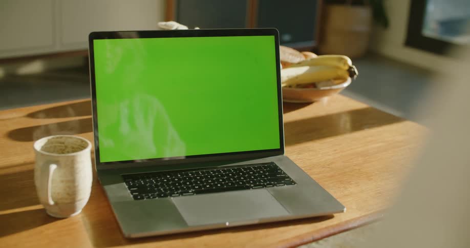 A laptop sits on a table in a home next to a mug with a steaming beverage in it